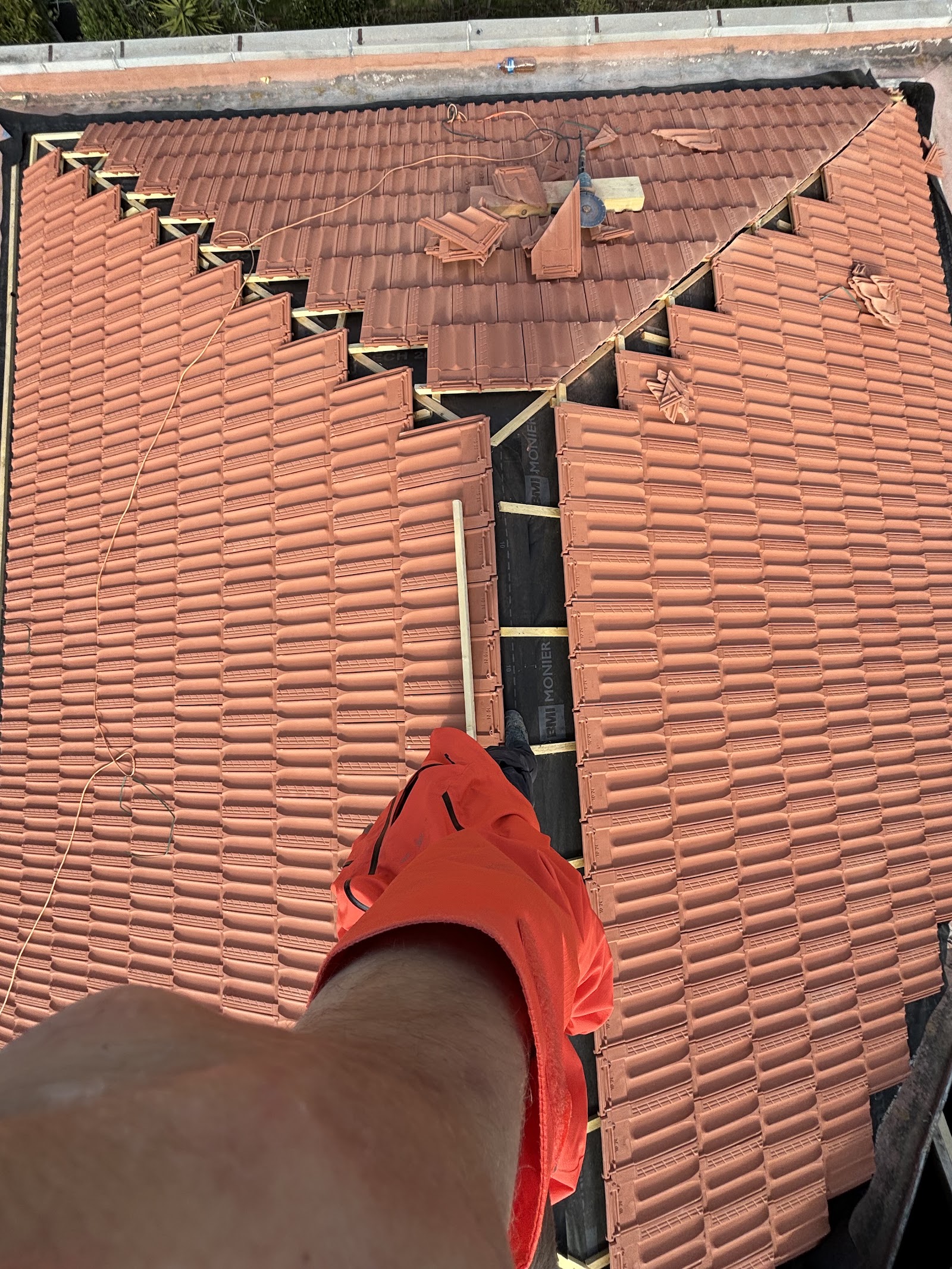 Vue en plongée sur le faîtage d'une toiture en tuiles, meuleuse et chutes de tuiles posées sur la couverture, bras d'un ouvrier au premier plan