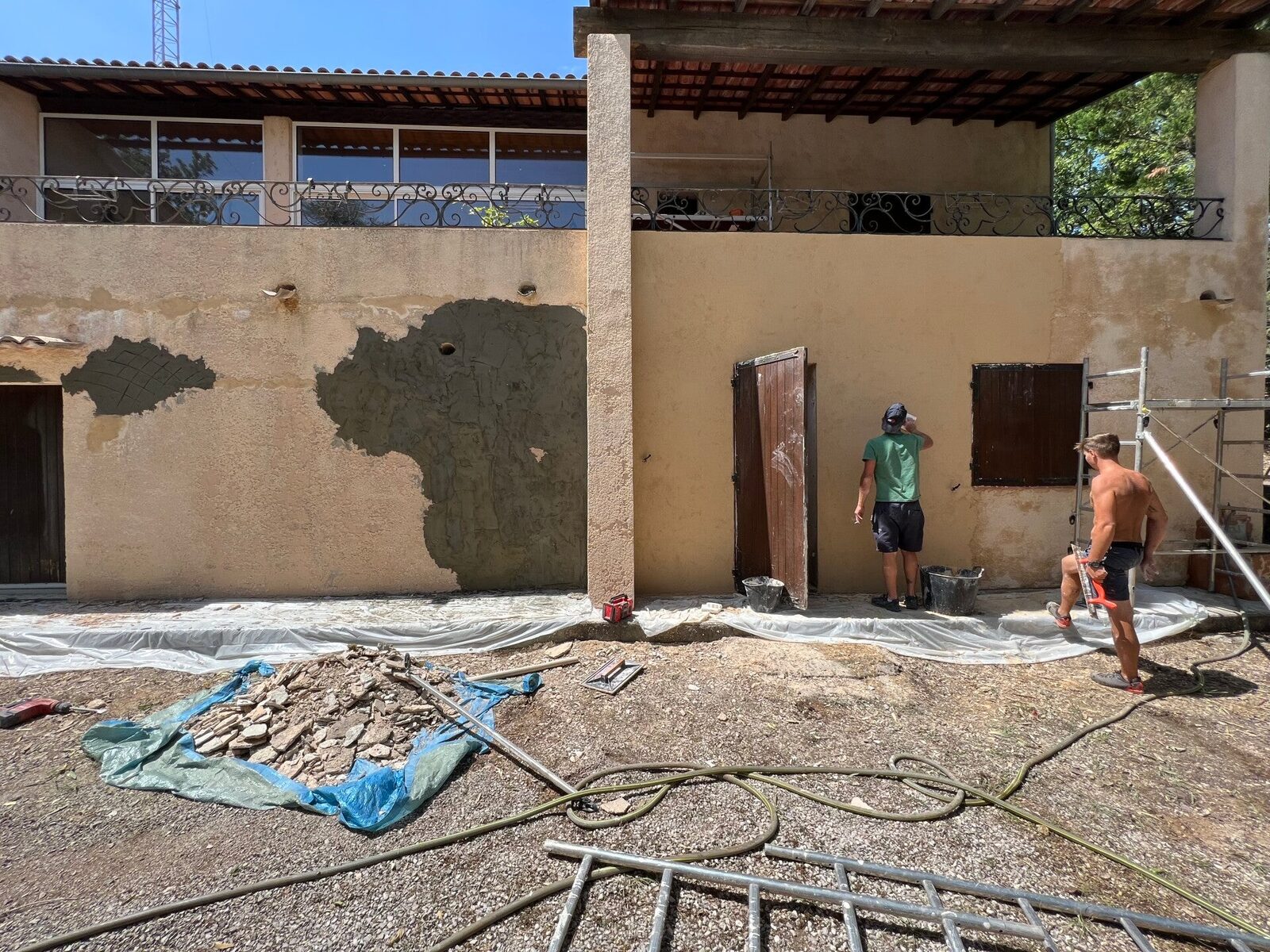 Ouvriers sur une façade avec zones d'enduit à nu et enduit frais appliqué, bâches de protection au sol